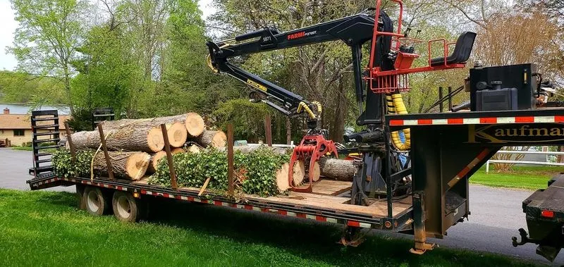 A trailer-mounted grapple loader stacked with cut logs and brush, parked on a driveway ready to haul off