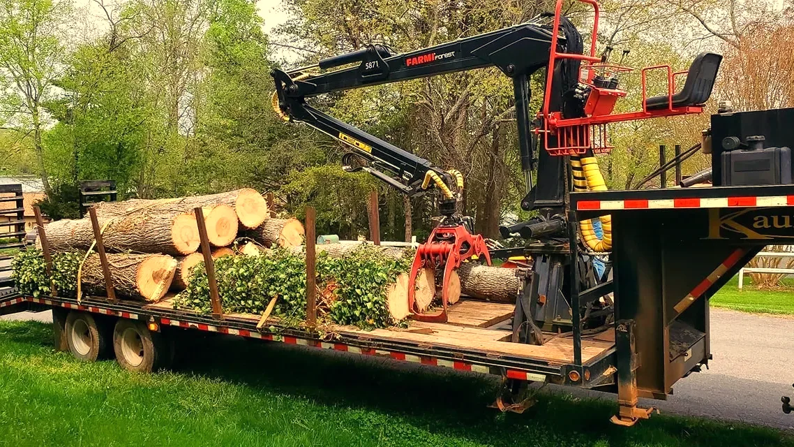 A truck-mounted knuckleboom crane with its grapple closed on a cut log, loading onto a trailer with its stabiliser legs down on the grass.